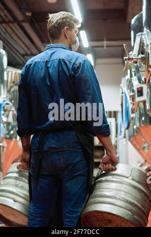 Vue arrière de l'ouvrier masculin de l'usine de production de bière moderne tenir des citernes entre deux rangées d'équipements de traitement Banque D'Images