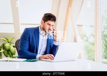 Photo d'un homme d'affaires sérieux utilisant son téléphone portable et parlant avec quelqu'un tout en étant assis au bureau et woking sur l'ordinateur. Banque D'Images