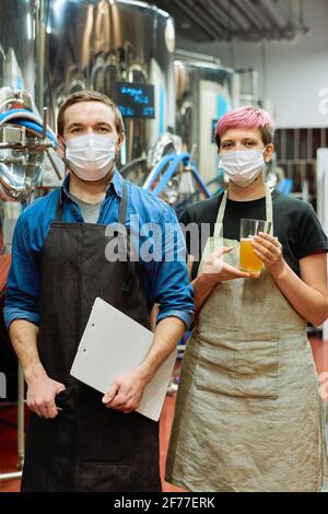 Deux jeunes professionnels de brasserie confiants dans les masques de protection debout avant de la caméra à l'intérieur de l'atelier de production de bière Banque D'Images