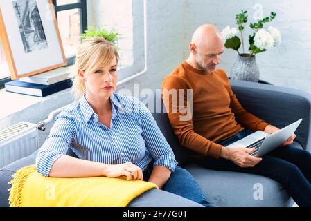 Prise de vue d'un couple d'âge moyen ignorant l'un l'autre après avoir un argument sur le canapé à la maison. Femme blonde assise sur le canapé pendant que son mari sitti Banque D'Images