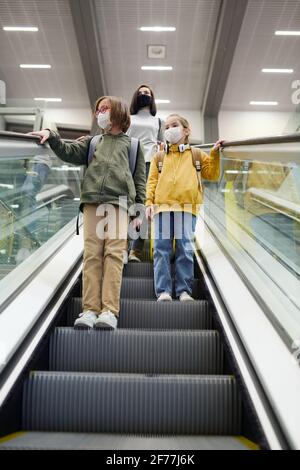 Deux jolies petites filles en vêtements décontractés et masques de protection et leur jeune mère debout sur les marches de l'escalier roulant dans moderne aéroport Banque D'Images