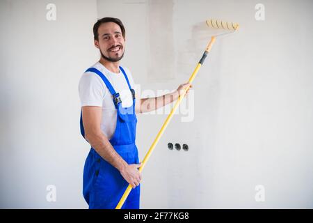 Un peintre barbu en vêtements de travail peint le mur avec un rouleau. Il sourit amical et pose pour la caméra Banque D'Images