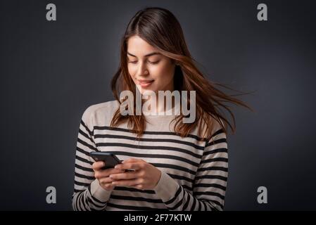 Portrait studio de femme souriante à l'aide d'un smartphone tout en se tenant sur un arrière-plan sombre isolé et en envoyant des messages texte. Banque D'Images