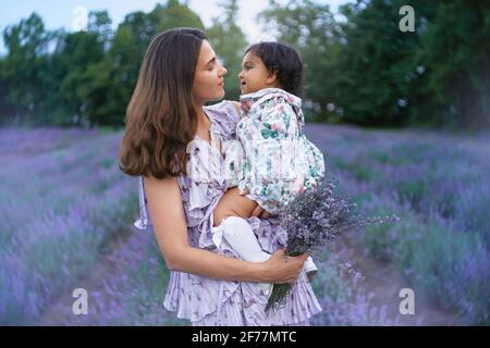 Jeune femme aimante posant avec bébé fille sur les mains dans le champ de lavande d'été. Vue latérale d'une mère heureuse portant une robe portant un joli bouquet de fleurs violettes. Concept de beauté de la nature. Banque D'Images