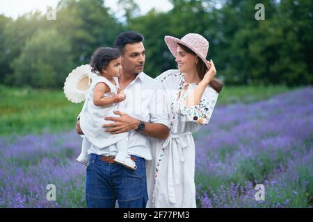 Famille heureuse mère, père et fille posant dans le champ de lavande. Petite fille de bébé portant chapeau de paille assis sur les mains de papa aimant, regardant maman à l'extérieur, fleurs aromatiques. Famille, concept de nature. Banque D'Images