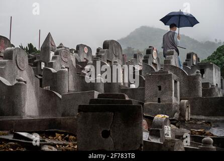 Un homme portant un parapluie marche à travers des pierres tombales dans un cimetière pendant le Ching Ming Festival à Hong Kong. Le Ching Ming Festival annuel, quand les gens visitent les tombes de parents décédés et quittent les lieux en souvenir et dans le respect que le gouvernement de Hong Kong tente de garder le nombre de cas infectés par le coronavirus COVID-19 sous contrôle tandis que davantage de personnes sont vaccinées. Banque D'Images