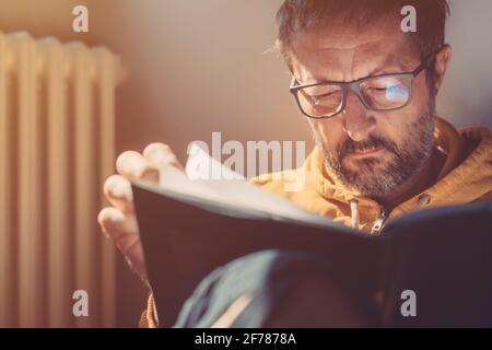 Homme pensif d'adulte moyen lisant un livre à la maison, portrait de gros plan de tête avec mise au point sélective, lumière du soleil brillant de la fenêtre Banque D'Images