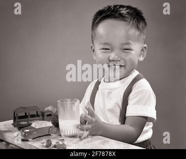 JEUNE GARÇON CHINOIS DES ANNÉES 1950 SOURIANT TENANT UN VERRE DE LAIT À LA CAMÉRA JOUANT AVEC LA VOITURE ET LE CAMION JOUET - C4688 HAR001 HARS DEMI-LONGUEUR HOMMES B&W CONTACT VISUEL GAI BOISSON ORIENTAL ET FLUIDE ASIATIQUE AMÉRICAIN DE SOURIRES CONSOMMER HYDRATATION JOYEUSE ASIATIQUE-AMÉRICAIN JUVÉNILES BOISSONS RAFRAÎCHISSANTES AUX PROTÉINES NOIR ET BLANC HAR001 À L'ANCIENNE Banque D'Images