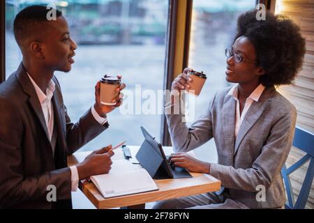 Souriant afro américain homme et femme buvant du café frais au café tout en dirigeant la réunion d'affaires. Deux collègues en tenue formelle utilisant des gadgets modernes pour le travail. Banque D'Images