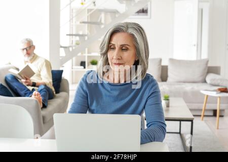 Femme mature travaillant en ligne depuis la maison tandis que le vieux mari se détend dans le salon. Banque D'Images