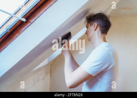 Un jeune homme en grattant avec une spatule et en enlevant le vieux papier peint sur un mur d'une pièce. C'est à vous. Rénovation de la maison. Mise au point sélective, copie Banque D'Images
