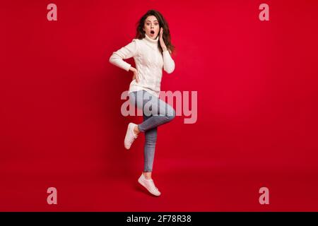 Pleine photo de la jolie jeune femme impressionnée vêtue de blanc pull sautant haut bras joue isolé rouge arrière-plan Banque D'Images