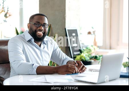 Homme d'affaires afro-américain, agent immobilier ou pdg, à la belle et confiante, vêtu de vêtements et de lunettes élégants, assis sur le lieu de travail, regardant un appareil photo, souriant et amical Banque D'Images