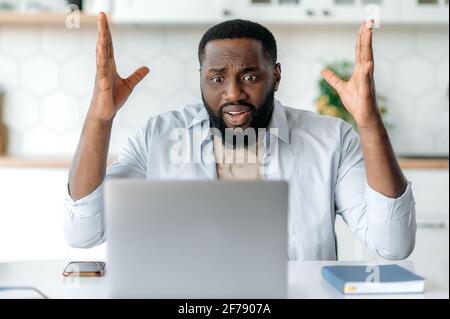 Choqué, confus, triste mécontent, un homme barbu afro-américain, découragé de regarder un ordinateur portable, a reçu un message inattendu ou des nouvelles, en faisant des gestes avec ses mains, se trouve sur le lieu de travail Banque D'Images