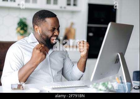 Joyeux confiant jeune africain-américain freelancer, Manager ou courtier, vêtu de vêtements élégants, travailler à distance, célébrer le succès, la victoire ou le bon profit, gesturant avec ses mains, souriant heureux Banque D'Images
