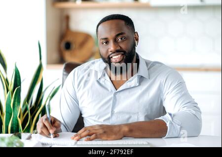 Tête de photo d'un homme barbu afro-américain joyeux dirigeant d'affaires, directeur, avocat ou courtier, portant une chemise bleue formelle, assis au bureau, prenant des notes, regardant l'appareil photo, souriant amical Banque D'Images