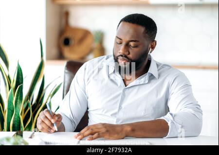 Concentré réussi confiant jeune Africain américain barbu, chef d'entreprise, directeur ou avocat travaillant ou étudiant à distance, dans une tenue formelle, assis à un bureau et prenant des notes focalisées Banque D'Images