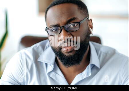 Portrait en gros plan d'un élégant homme barbu afro-américain sérieux et sûr de succès avec des lunettes, portant une élégante chemise habillée, concentrée à regarder l'appareil photo Banque D'Images