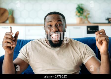 Joyeux joyeux jeune Africain américain barbu, dans des vêtements élégants, assis sur le canapé, tenant un smartphone et une carte de crédit, en faisant des gestes avec ses mains, souriant heureusement, a obtenu une réduction pour faire du shopping Banque D'Images
