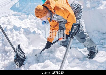 Un jeune homme de race blanche en costume d'hiver jaune tient une pelle à neige. Un garçon nettoie l'arrière-cour de la neige. Jour ensoleillé, froid glacial, acti en plein air Banque D'Images