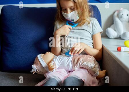 Une petite fille en masque médical est assise sur le canapé, joue un médecin, traite et écoute la poupée avec un stéthoscope dans un masque médical, à la maison pendant qu Banque D'Images