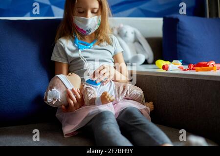 Une petite fille en masque médical est assise sur le canapé, joue un médecin, traite à la poupée Banque D'Images