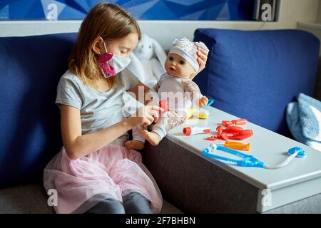 Une petite fille en masque médical est assise sur le canapé, joue un médecin, traite à la poupée Banque D'Images