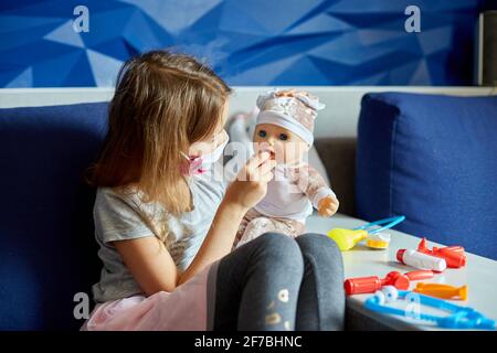 Une petite fille en masque médical est assise sur le canapé, joue un médecin, traite à la poupée Banque D'Images