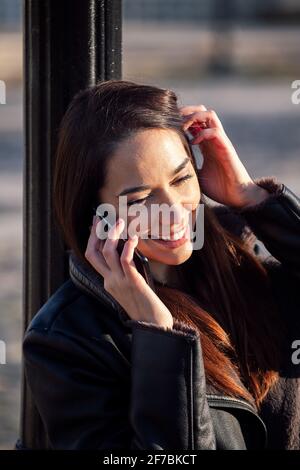 photo verticale d'une jeune femme souriante qui parle au téléphone et qui se penche contre un lampadaire, le concept de technologie et le mode de vie urbain Banque D'Images