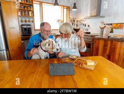 Couple senior heureux avec un chien et du vin en dégustant des vidéos en ligne avec des amis et des membres de la famille célébrant à distance les vacances d'hiver. Coronavirus second wa Banque D'Images