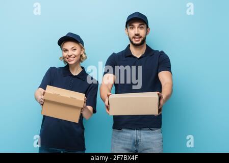des coursiers interraciaux positifs montrant des boîtes en carton tout en regardant l'appareil photo isolé sur bleu Banque D'Images
