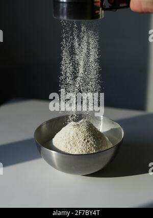Main de l'homme chef tamisant la farine à travers le sifter en acier sur le tableau blanc. Concept de préparation de biscuits, de cuisson de tarte ou de pâtisserie de Pâques. Photographie verticale. Image de haute qualité Banque D'Images