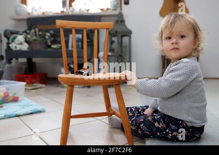 kleines Mädchen sitzt im Wohnzimmer auf dem Boden neben einem Kinderstuhl aus Holz - petite fille est assise sur le sol à côté d'une chaise d'enfant en bois dans le livi Banque D'Images