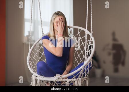 Une belle femme blonde en vêtements de sport bleus, pratiquant le yoga, est engagée dans la méditation dans la position lotus, assis dans une chaise suspendue dans la salle, Banque D'Images