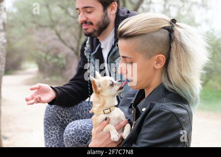 Chihuahua léchant ses propriétaires bouche et ami dégoûté à côté. Jeune couple punk dans un parc. Banque D'Images