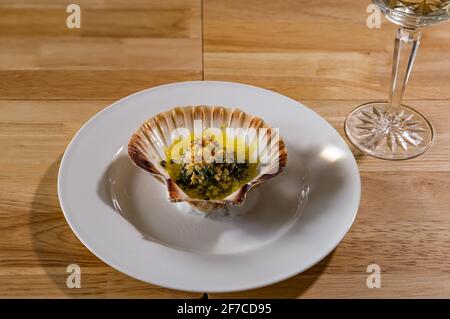 Menu dégustation plat de poisson : pétoncles au beurre d'ail, câpres et miettes de parmesan servis dans des coquilles de pétoncles sur une assiette blanche et un verre cristal Banque D'Images