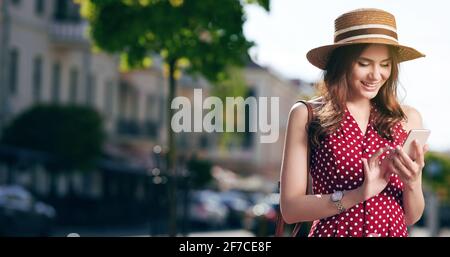 Une femme caucasienne écrit des messages sur son smartphone et entre la ville d'été avec un chapeau de paille et un beau robe rouge à pois Banque D'Images