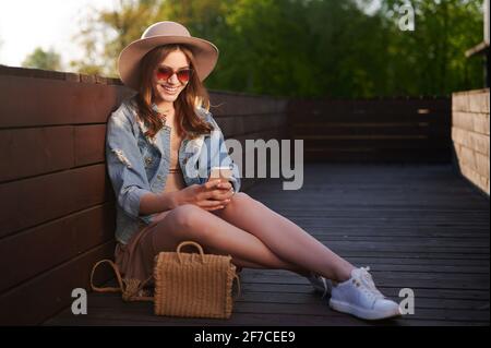 Femme souriante dans la tenue d'été chapeau de paille utilise un smartphone dehors et assis sur la terrasse en bois Banque D'Images