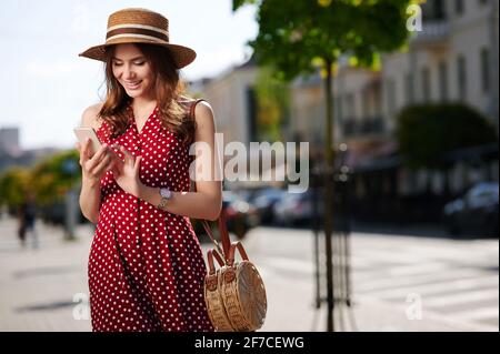 Belle jeune femme utilisant un smartphone et des promenades en été ville portant chapeau de paille et belle robe rouge avec polka points Banque D'Images