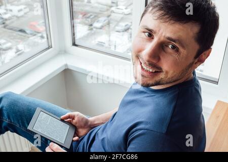Souriant beau barbu homme relaxant dans un bleu jeans et t-shirt lisant un livre électronique sur un appareil numérique du balcon près de la fenêtre dans un soupir Banque D'Images