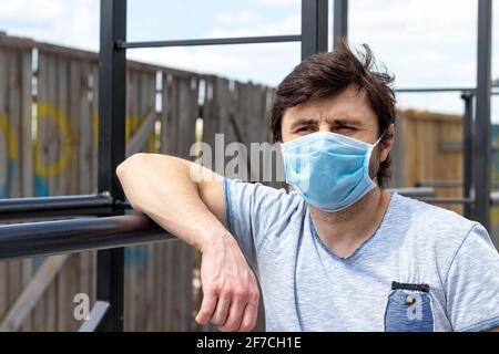 Homme dans un masque médical bleu de protection debout sur le terrain de sport en plein air, reposant sa main sur les barres. Homme dans un respirateur pour protéger contre les infaux Banque D'Images