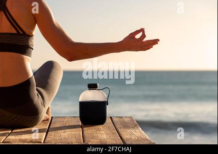Gros plan d'une femme qui fait de la méditation au bord de la mer au lever ou au coucher du soleil avec une bouteille d'eau pour rester hydraté. Banque D'Images