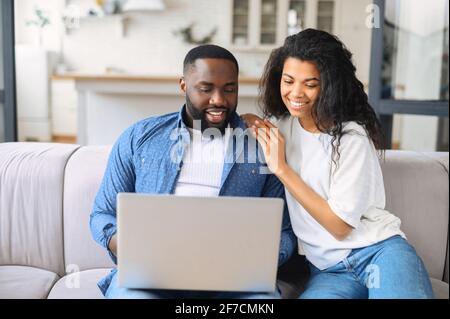 Jeune couple afro-américain noir utilisant un ordinateur portable assis sur le canapé à la maison. Couple pleine longueur regardant un film sur un ordinateur portable. Un homme et une femme heureux se détendent sur un canapé et passent du temps libre à la maison Banque D'Images