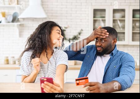 Black man a vu une facture pour les achats en ligne de sa femme tenant la carte de crédit, femme hispanique avec le téléphone mobile, heureux après avoir acheté des vêtements ou un sac coûteux, le gars anxieux garde sa main sur le front Banque D'Images