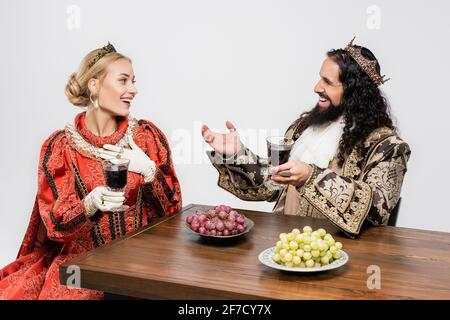 joyeux couple historique interracial dans les couronnes tenant des verres de rouge vin assis à table avec des raisins isolés sur du blanc Banque D'Images