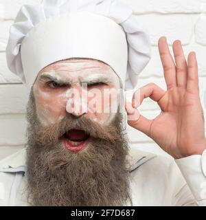 L'homme avec de la farine sur le visage montre OK. Gros plan de boulanger sérieux, cuire avec de la farine sur le visage, la barbe et la moustache poses en uniforme de chef et chapeau Banque D'Images