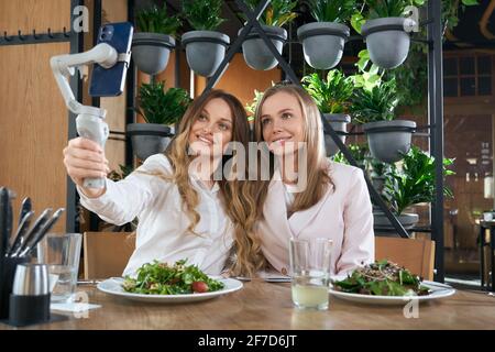 Vue de face de jeunes femmes souriantes blogger assis à la table avec de savoureuses salades dans le café moderne et prendre un selfie. Concept de dîner et de communication avec bonne humeur. Banque D'Images