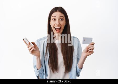 Femme caucasienne excitée montrant la carte de crédit et tenant le smartphone, faisant la commande, attendant la livraison tout en faisant des achats en ligne, debout sur blanc Banque D'Images