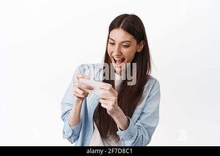 Une jeune fille excitée jouant à un jeu vidéo de course sur un smartphone, jouant sur un téléphone mobile, regarde l'écran joyeuse et souriante, se tenir sur un fond blanc Banque D'Images