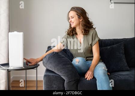 La technologie pour une vie domestique saine. Jeune femme qui active l'humidificateur pour l'humidification de l'air, le nettoyage et le rafraîchissement, femme de ménage souriante assise sur le canapé Banque D'Images